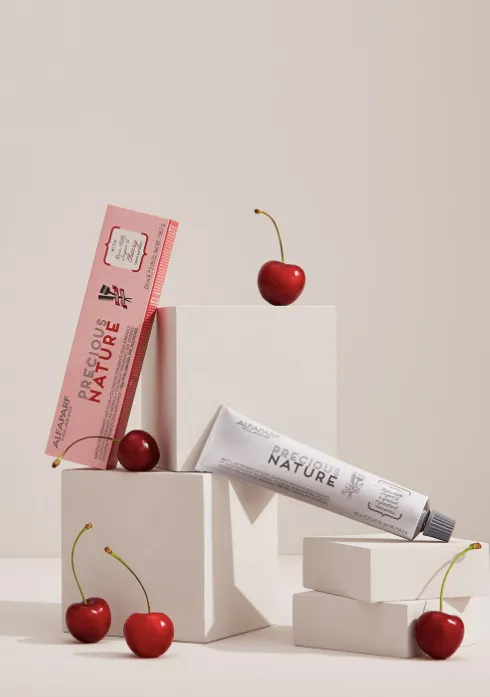 Tinte capilar Alfaparf Precious Nature con cerezas, presentación del producto en tubo y caja sobre bloques blancos minimalistas