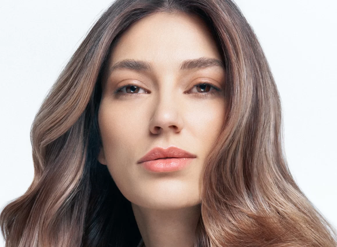 Primer plano de mujer con cabello largo castaño con ondas suaves y mechas bronde balayage, blusa satinada beige Alfaparf tonos perlados