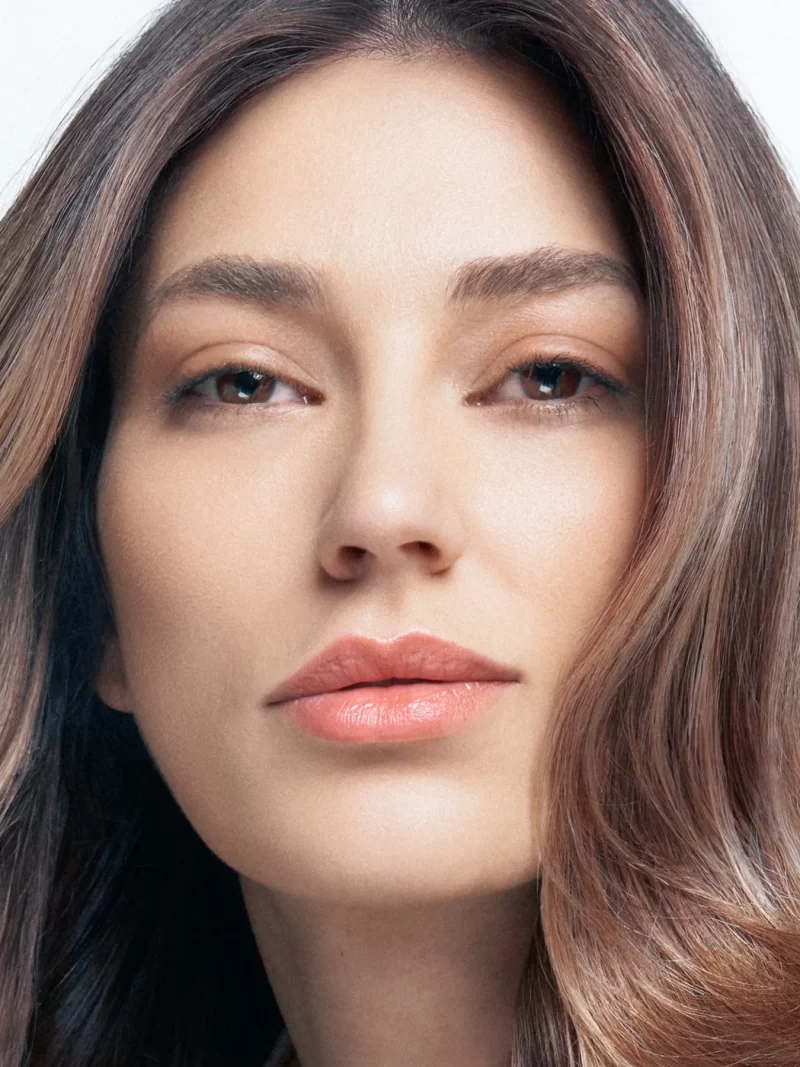 Primer plano de mujer con cabello largo castaño con ondas suaves y mechas bronde balayage, blusa satinada beige Alfaparf tonos perlados