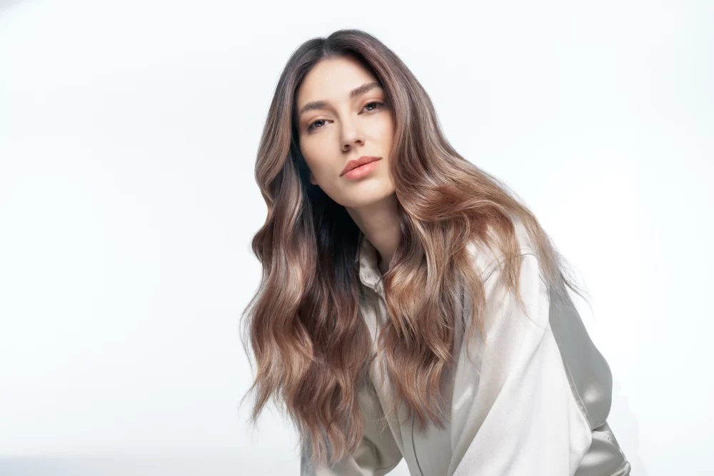 Primer plano de mujer con cabello largo castaño con ondas suaves y mechas bronde balayage, blusa satinada beige Alfaparf tonos perlados