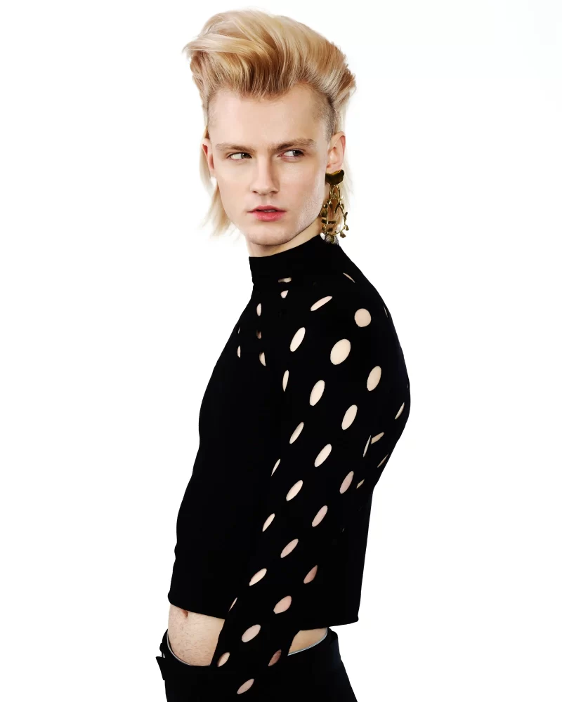 Modelo masculino con mohawk rubio alto, blusa negra con cortes circulares y arete colgante dorado.