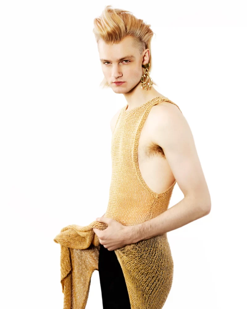 Mohawk rubio con raíces oscuras y vestido tejido dorado, estilo fluido con acento dramático.