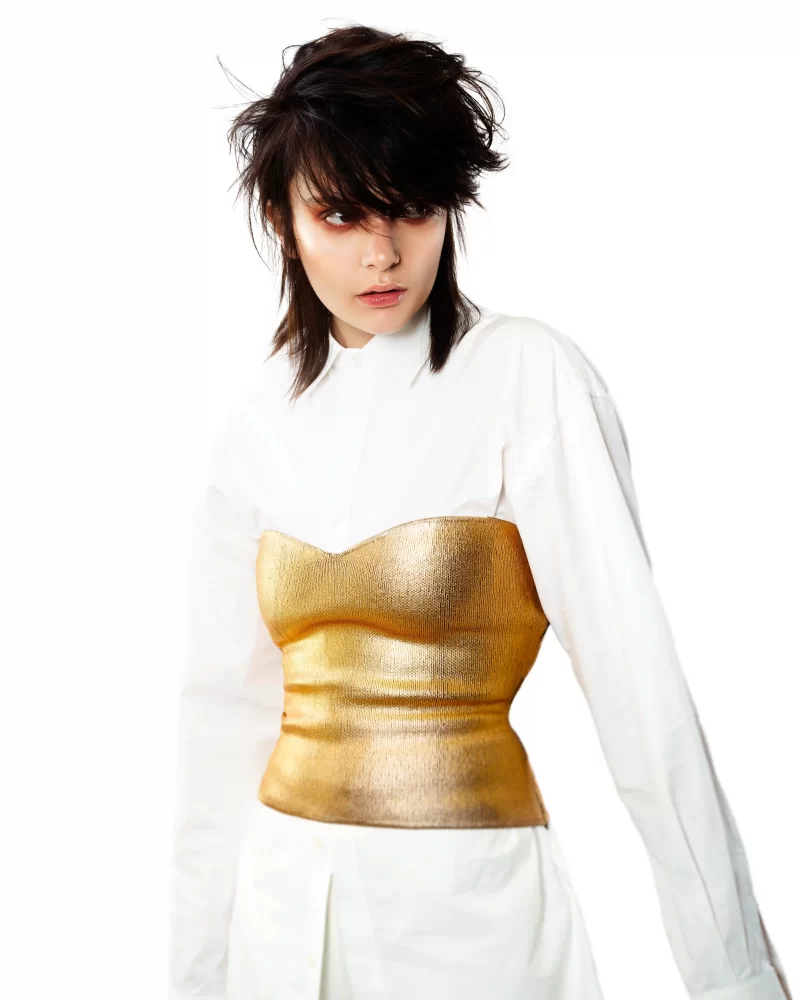 Peinado shag oscuro con flequillo desordenado, camisa blanca y corset dorado, estilismo queer sin etiquetas.