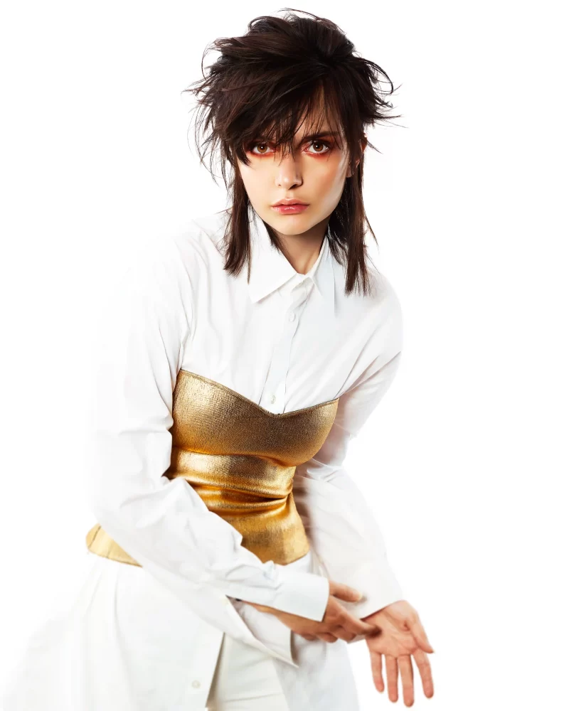 Modelo con corte shag oscuro, camisa blanca y corset dorado metálico, mirada intensa y postura editorial.