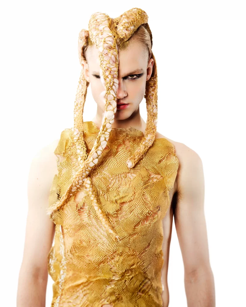 Peinado escultórico con trenzas doradas sobre el rostro y top de malla dorada, estética tribal futurista.