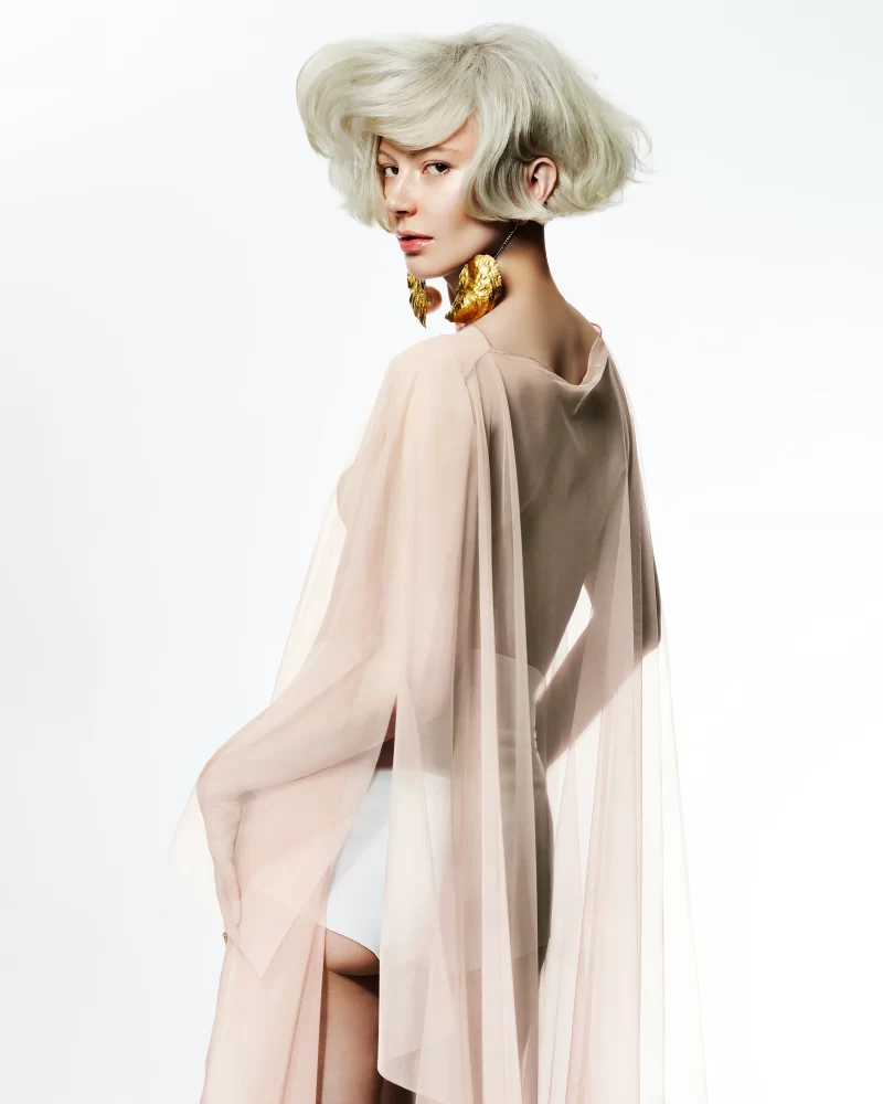 Modelo con peinado ondulado rubio platinado, vestido nude translúcido y aretes dorados en forma de hojas.