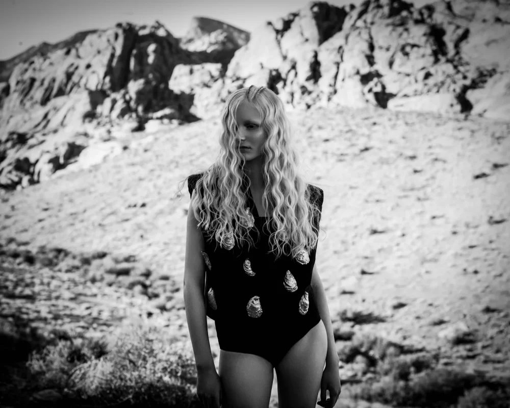 Modelo rubia con ondas largas y marcadas, vestida con body negro decorado, en paisaje desértico en blanco y negro.