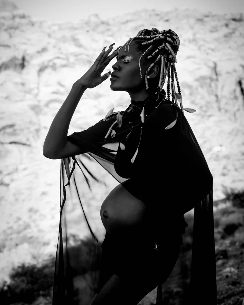 Modelo embarazada con peinado trenzado alto y accesorios étnicos, en entorno desértico en blanco y negro