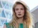 Modelo rubia con cabello suelto y textura ligera, vistiendo blusa verde frente a edificio contemporáneo de cristal.