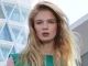 Modelo rubia con cabello suelto y textura ligera, vistiendo blusa verde frente a edificio contemporáneo de cristal.