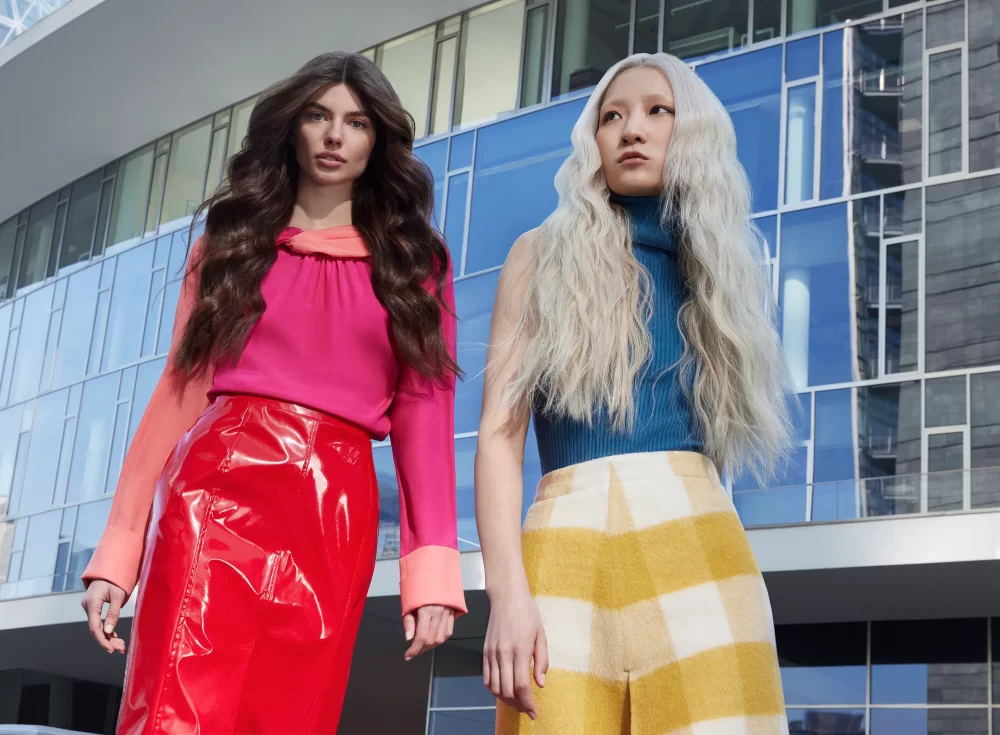 Dos modelos con cabello largo, una con melena oscura en ondas marcadas y otra con cabello platinado y suelto, posando frente a edificio de vidrio azul.