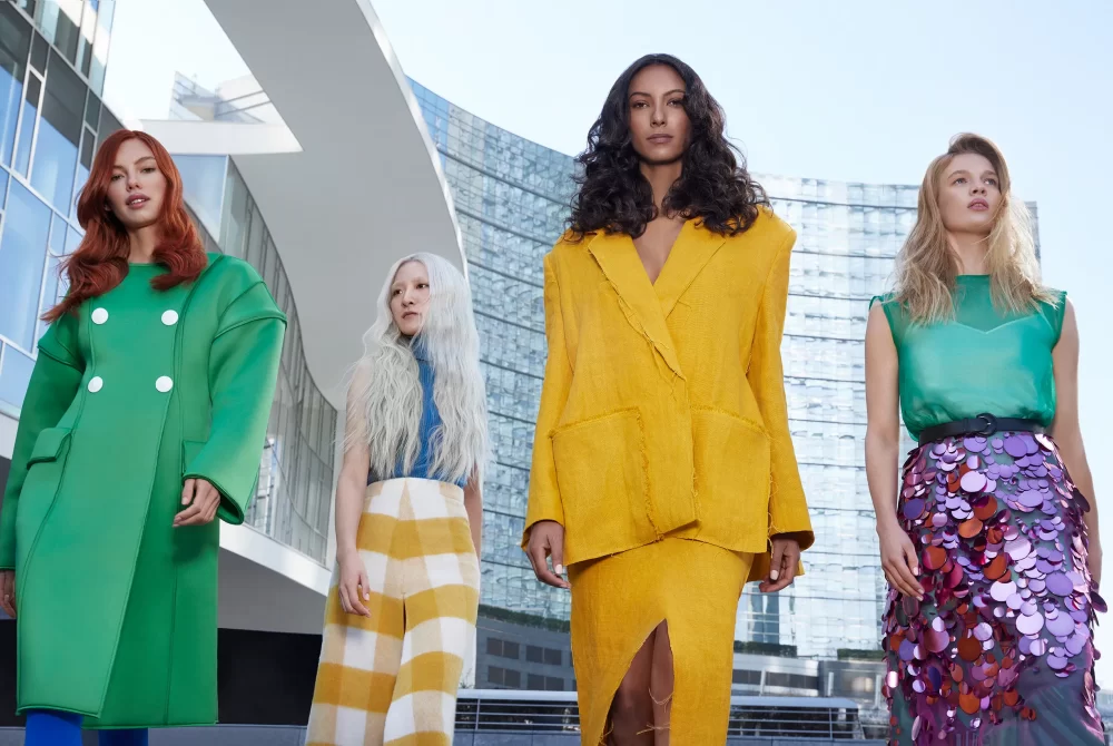 Grupo de cuatro modelos con distintos tonos y texturas de cabello, vestidas con outfits coloridos frente a arquitectura moderna de cristal.