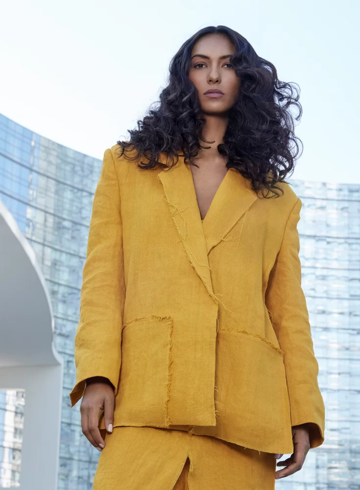 Modelo afrodescendiente con melena afro voluminosa en tonos cobrizos, usando vestido metálico naranja frente a fondo arquitectónico blanco.