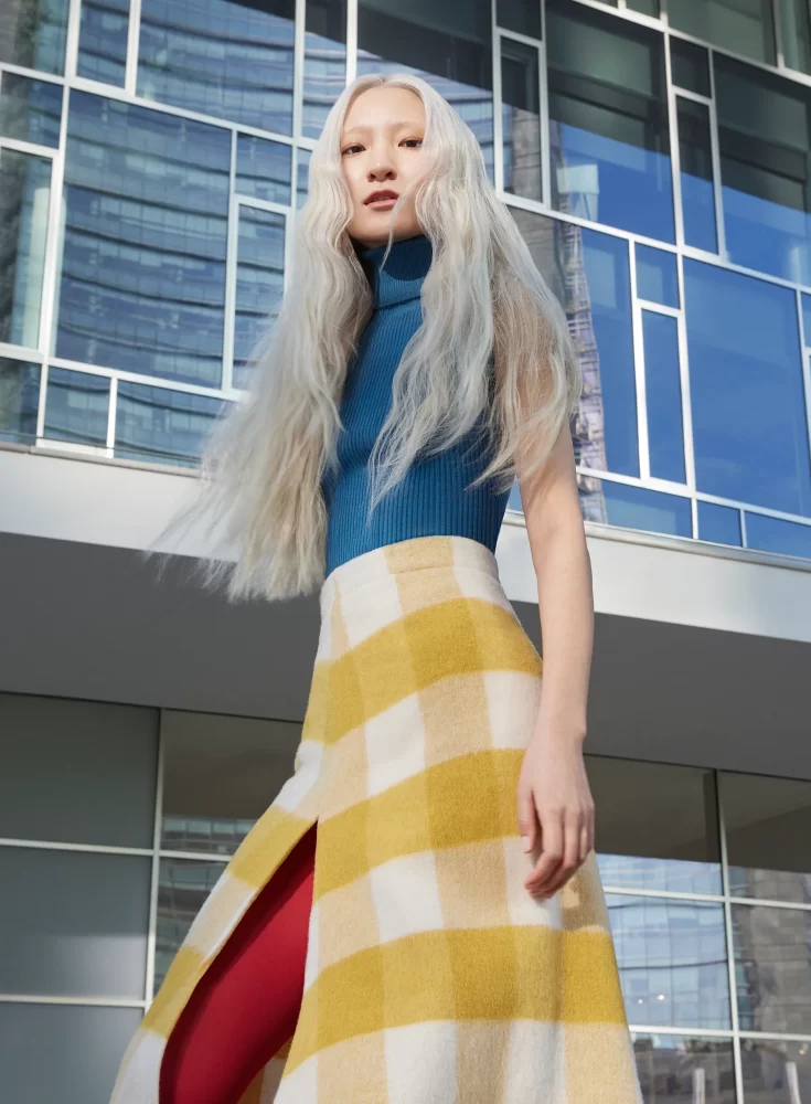 Modelo asiática con cabello largo negro intenso y flequillo recto, vestida de blanco frente a fondo arquitectónico moderno y neutro.