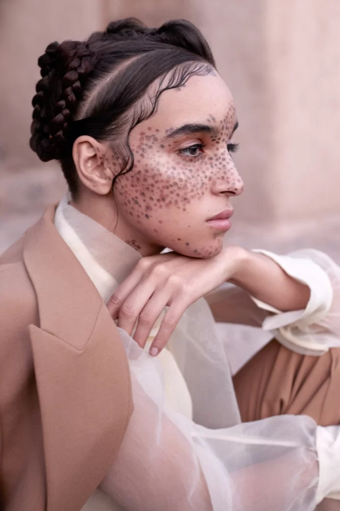Modelo de perfil con maquillaje punteado artístico, peinado trenzado y blazer beige de la colección Divine Feminine de Juliette Den Ouden.