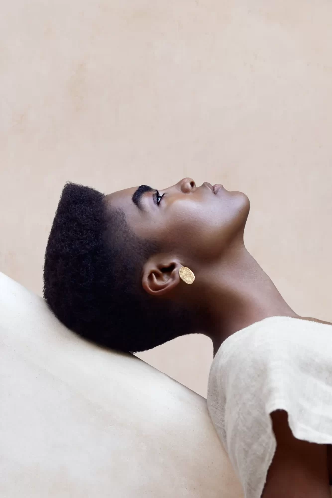 Modelo de piel oscura recostada en bloque blanco, con afro flat top y pendiente dorado de Divine Feminine por Juliette Den Ouden.
