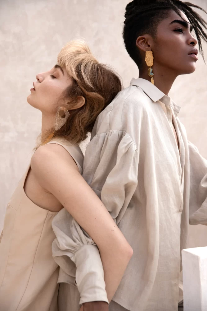 Dos modelos de espaldas unidas, una de piel clara con cabello rubio y otra de piel oscura con dreadlocks y pendientes dorados, de Divine Feminine por Juliette Den Ouden