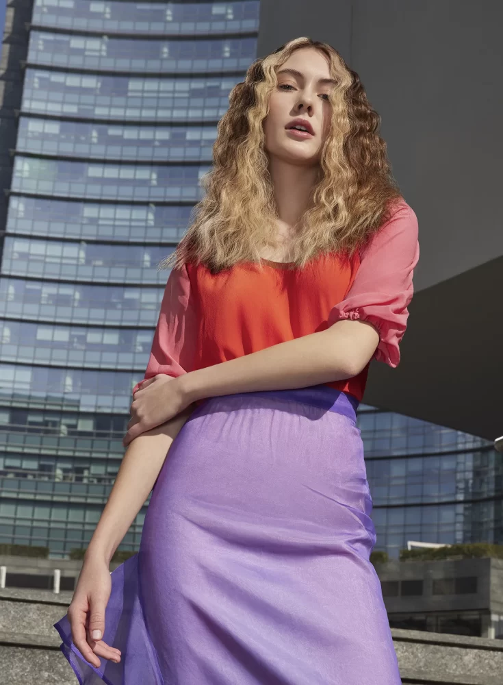 Modelo rubia con melena media en ondas suaves y vestido metálico lila frente a pared de concreto claro.