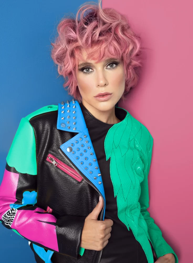 Modelo con cabello rizado en tono rosa pastel, usando chaqueta con apliques verdes y azul eléctrico. Colección Metaverso de Alexander Kiryliuk.